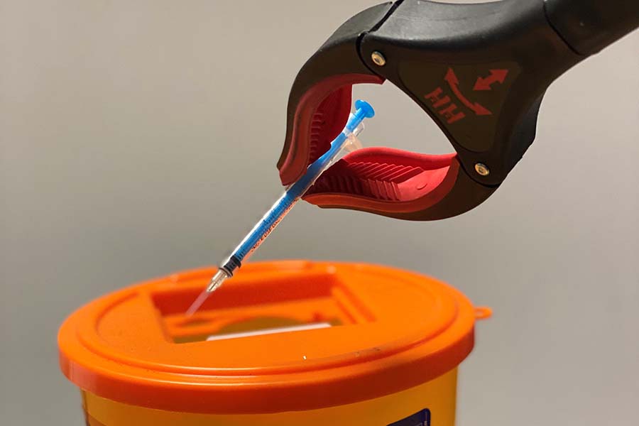 A hypodermic needle being gripped by a sharps picker and placed into a disposal bin